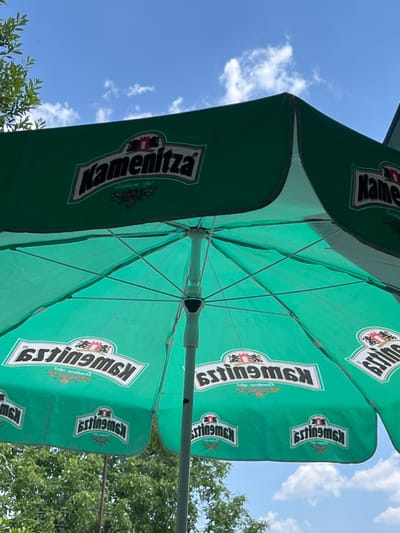 A vibrant green outdoor umbrella, likely a patio umbrella for a restaurant or cafe, dominates the frame. The umbrella is open and tilted upwards, revealing its internal structure of thin metal poles. The fabric of the umbrella is emblazoned with logos and text. On one panel, the word "Kamenitza" is clearly visible in a stylized font, suggesting the umbrella is a branded item. Other panels feature the text "Kswu9msj" and some smaller script text, along with emblems containing what appear to be crests or decorative insignias.

The umbrella is set against a bright blue sky with scattered white clouds, indicating fair weather and likely daytime, possibly late morning or early afternoon given the brightness. Lush green foliage, likely from trees, is visible in the background and along the bottom edge of the image, suggesting an outdoor setting in a natural or park-like environment. The overall impression is one of a sunny, pleasant day, suitable for outdoor leisure activities. The orientation of the image, looking up at the umbrella from below, emphasizes its scale and design. The location context provided, Branipole, Bulgaria, adds a specific geographical element to the scene.