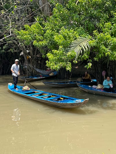 The image shows several small blue boats on a murky, greenish-brown river, possibly a canal, in Tân Thạch, Vietnam. A man stands barefoot in the foremost boat, wearing a white shirt, dark pants, and a white cap, holding a long pole, likely used for maneuvering the boat. Other boats contain seated individuals, some wearing traditional conical hats. Lush green vegetation lines the riverbank.