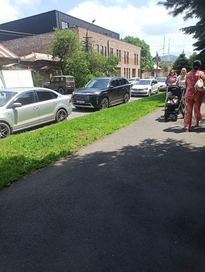 In Atabe, Georgia, a paved walkway runs parallel to a street lined with parked cars and a grassy verge. On the walkway, a person pushes a stroller with a baby. Further down the street, several cars are parked, including a silver sedan and a black SUV. Behind the cars, a modern brick building with a dark metal roof stands against a backdrop of trees and a clear sky. The scene depicts a casual urban environment on a sunny day.