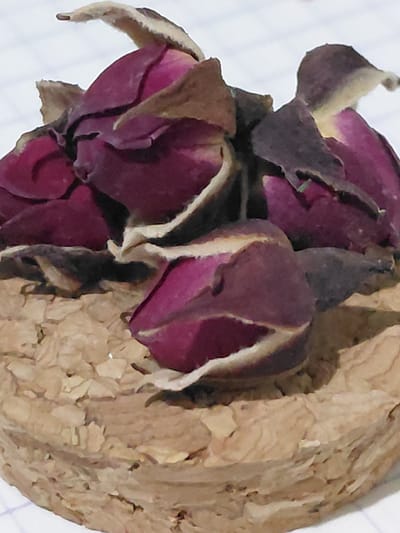 The image shows a close-up, macro view of several dried rose buds. The rose buds are a deep magenta color with visible darker brown and tan dried petals and sepals. They are clustered together and appear to be resting on a textured, light brown material that resembles dried plant matter or a natural fiber. The background is a blurry white surface with faint grid lines, suggesting it might be paper. The lighting is diffused, with some shadows highlighting the texture of the roses and the base material. The image is taken from a slightly elevated angle, focusing on the arrangement of the rose buds. There are no people or any indication of a specific location in the city of Ертіс ауылдық округі, Kazakhstan.