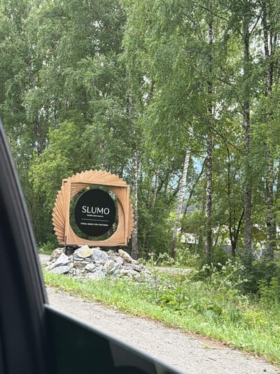 The image shows a wooden sign with the text "SLUMO hotels and resorts" and Russian text below it, placed on a pile of rocks. The sign is set against a backdrop of lush green trees. A dark car window frame is visible in the foreground, suggesting the photo was taken from inside a vehicle. The surrounding environment is natural and wooded, with a gravel road leading past the sign.