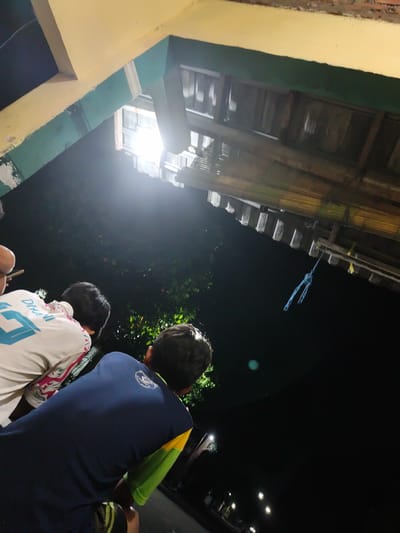 In Barengkrajan, Indonesia, at night, three individuals are seen from behind, looking up towards a structure. The structure appears to be a roof or overhang with a yellow and green painted beam. Below this structure, a wooden roof with bamboo elements is visible, and a blue rope is hanging from it. The background is dark, indicating nighttime, with some distant streetlights and foliage. One person in the foreground is wearing a dark blue t-shirt with green and yellow accents. Another person to their left is wearing a white t-shirt with blue lettering and designs. The third person, partially visible, is in the center. The lighting suggests an outdoor setting, possibly a gathering or observing an event.