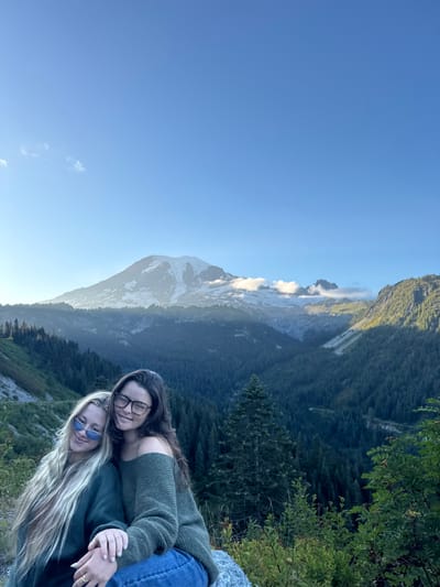 Two young adult females are seated on natural ground in the foreground of a mountainous landscape. The individual on the left has long blonde hair, wears blue sunglasses, and a dark green long-sleeved top. The individual on the right has long dark hair, wears clear-frame eyeglasses, an olive green off-the-shoulder sweater, and blue jeans. Her left arm is positioned around the left individual, and her left hand rests on the left individual's right hand. Both subjects are looking towards the camera and smiling.
The background features a prominent, snow-capped stratovolcano identified as Mount Rainier, under a clear blue sky with minimal cloud cover. The midground consists of dense coniferous forests filling valleys and covering lower mountain slopes. The foreground terrain is rocky with low-lying green and reddish-brown vegetation. Illumination indicates a daytime setting, likely early morning or late afternoon. The scene is located in a mountainous region of the United States, consistent with an elevated viewpoint within or bordering Mount Rainier National Park, which is geographically accessible from the city of Packwood, Washington.