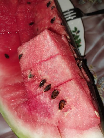 The image shows a close-up of a sliced watermelon, with its vibrant pink flesh and dark seeds prominently displayed. The watermelon is cut into several pieces, with one large chunk dominating the foreground. The texture of the watermelon is glistening and juicy, with small droplets of moisture visible on its surface. Several dark, oval-shaped seeds are scattered across the flesh, some partially embedded.
In the background, a blurry dark object, likely a fork, rests on a plate adorned with a floral pattern in green and blue. The plate is placed on a surface covered with a patterned fabric, possibly a tablecloth, with colors of pink and brown. The overall impression is a still life of a freshly cut watermelon, suggesting a refreshing treat. There is no indication of the time of day, weather conditions, or specific emotions. The location context of Rohati Community of villages in Tajikistan is not visually evident in the image itself. No visible text is present.