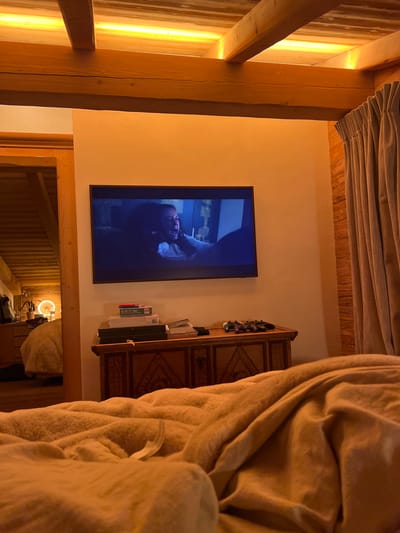This image depicts the interior of a residential space, characteristic of a chalet-style dwelling, inferred to be in Saanen, Switzerland. The perspective is low, consistent with a view from a bed in the foreground, covered with rumpled, light-colored bedding.
A flat-screen television is mounted on a light-colored central wall. The screen displays a dark, intense scene featuring a person, likely female, with dark hair, screaming or yelling, their mouth agape, with what appears to be another hand near their face.
Below the television, a wooden chest or low cabinet with decorative carvings holds a stack of items, including a box faintly labeled "DO NOT DISTURB," and several video game controllers on its right side.
The ceiling features prominent exposed wooden beams with concealed LED strip lighting casting a warm, upward glow. To the right, a wall with horizontal wooden paneling is partially obscured by a light-colored, pleated curtain. A wooden door frame on the left leads into an adjacent room, where another bed with light-colored bedding, a bright circular lamp, and indistinct objects are visible in the background. The overall lighting is warm and artificial. No individuals are directly visible in the depicted rooms.