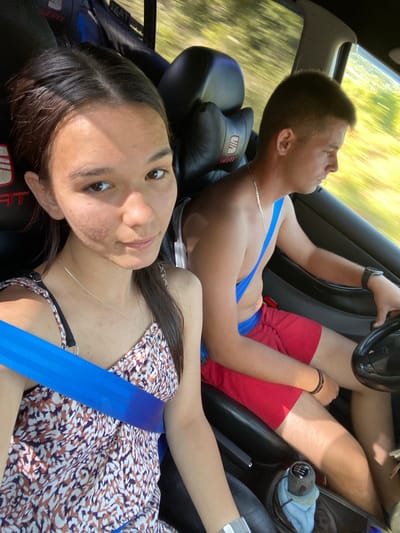 Two young people, a man and a woman, are inside a car in Berievo, Bulgaria. The man, in the foreground on the left, is driving. He is wearing red shorts and a silver chain. A blue seatbelt is across his chest. His face is turned away from the camera. He has short, dark hair and is wearing a watch on his left wrist.
The woman is in the passenger seat on the right, leaning back and looking directly at the camera. She has long, dark hair and is wearing a patterned dress. A blue seatbelt is across her chest and shoulder. Her expression appears relaxed or amused.
The scene is likely taking place during the daytime, with sunlight filtering through the car windows, casting bright light on the scenery outside. The background visible through the windows shows greenery, suggesting a rural or semi-rural location. The car interior is visible, including the steering wheel, gear stick, and seats. The overall atmosphere seems casual and perhaps a bit playful, given the woman's direct gaze at the camera. The location cue "Berievo, Bulgaria" is provided. No visible text is apparent in the image.
