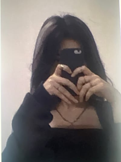The image depicts an individual taking a selfie in Ittenheim, France. The person's face is obscured by a dark-colored smartphone held in both hands. The fingers of both hands are positioned to form a heart shape in front of the individual's upper chest/neck area. The person has long, dark hair and is wearing a dark, long-sleeved top. A thin necklace with a small pendant is visible around their neck, and a ring is present on the ring finger of the left hand. The background is a plain, light-colored, indistinct surface. The overall image quality is pixelated and blurry.