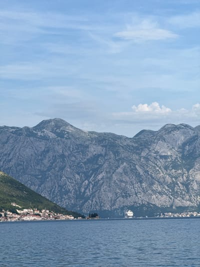 The image depicts a panoramic view of the Bay of Kotor in Montenegro, with a focus on the town of Kostanjica. The foreground is dominated by a calm, blue body of water, likely the bay itself. In the mid-ground, a coastal town is visible nestled at the base of steep, rocky mountains. The town is characterized by buildings with red-tiled roofs and white facades, indicative of Mediterranean architecture. To the right of the town, a small island with a few trees and buildings can be seen. Further out on the water, a large white cruise ship is present, moving from left to right. The background is composed of imposing, rugged mountains with a weathered texture, their slopes sparsely covered in vegetation. The sky above is a pale blue, with scattered white clouds. The overall impression is one of a serene coastal landscape with human settlements integrated into a dramatic natural environment.