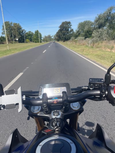 A motorcycle speeds down a straight, empty road under a clear blue sky. The dashboard displays a speed of 63 mph. Lush green trees line the road, illuminated by sunlight. The motorcycle's handlebars and controls are in sharp focus, contrasting with the blurred background.