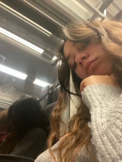 A close-up, angled photograph depicts a young female individual seated inside a public transport vehicle in Schwindratzheim, France. The subject, positioned in the right and central foreground, has long, wavy brown hair and is looking towards the camera with a neutral expression and slightly pursed lips. Her right hand, covered by a light grey ribbed knit sweater sleeve, is resting near her chin. The interior of the vehicle is visible in the background, featuring horizontal fluorescent strip lighting on the ceiling and a reflective window on the left, which shows indistinct dark shapes and reflections of the overhead lights. Dark seating is partially visible behind the primary subject.