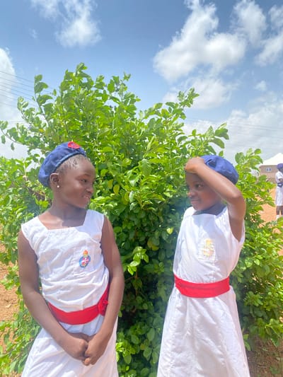 Two young people are dressed in white uniforms with red sashes, standing outside amidst green shrubs under a partly cloudy sky. Both are wearing navy blue caps. The location appears to be in Turu, Nigeria, though specific city details are not distinctly visible.