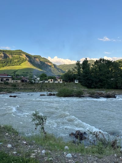 The image depicts a scenic view of the city of Каменномостское сельское поселение, Russia. A wide, fast-flowing river occupies the foreground, with choppy water and visible rocks. The riverbank is lined with gravel, stones, and sparse green vegetation, including a small, bare tree. In the middle ground, a cluster of houses and small structures are visible along the river. These buildings have red tiled roofs and are situated on a raised bank. Behind the settlement, a dense line of tall, dark green trees forms a treeline. In the background, rolling green hills rise sharply, with steep, rocky cliffs and grassy slopes. The sky is a clear, bright blue with a few scattered white clouds, suggesting daytime and fair weather. The overall scene conveys a sense of natural beauty and rural life in a mountainous region.