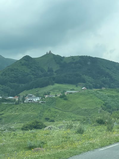 The image shows the Gergeti Trinity Church, a 14th-century Orthodox Christian church located in the city of Gergeti, Georgia. The church is situated on a steep hill, overlooking a valley with a small village below. The landscape is lush and green, with rolling hills covered in trees and wildflowers. The sky is overcast, with dramatic clouds adding to the atmospheric scene. In the foreground, a road is visible, suggesting the vantage point is from a vehicle or a roadside. Several houses and buildings are scattered across the lower slopes of the hills, forming the village. A flock of sheep is grazing in a pasture on the hillside. The overall impression is one of a remote, picturesque, and historically significant location.