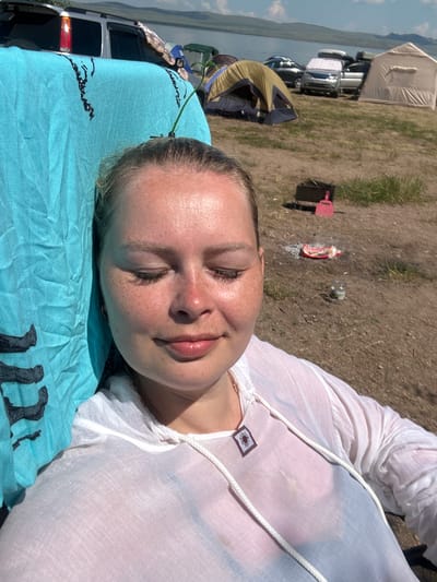 A young Caucasian woman with blonde hair, fair skin, and freckles is captured in a selfie at a campsite by a lake. She is leaning against a bright blue towel, with her eyes closed and a slight smile on her lips. She is wearing a sheer white hooded top over a dark-colored undergarment. Her hair is pulled back, and she has a small, square pendant with a sun symbol on it.
The background reveals a natural landscape with a body of water, hills, and a sunny sky with scattered clouds. Several tents and vehicles are visible, suggesting a group camping activity. A barbecue grill, a pink shovel, and some debris are scattered on the dry, grassy ground. The overall atmosphere is relaxed and informal, indicative of an outdoor recreational setting. The presence of tents and cars implies this is likely an established camping ground or designated recreational area.