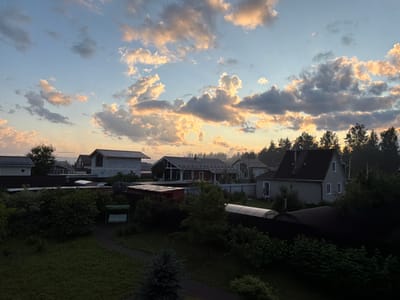 This media file captures a serene evening scene in Kerro, Russia. The sky is a canvas of soft blues and warm oranges, illuminated by the setting sun which casts a golden glow on scattered, fluffy clouds. Below the sky, a collection of houses with dark roofs and light-colored walls are nestled amongst lush green trees and manicured lawns. Some houses appear to be under construction, with exposed brickwork and unfinished roofs.

In the foreground, a winding path leads through a verdant garden, bordered by dense bushes and a small, dark-colored evergreen tree. A light-colored swing set with a striped canopy is visible on the path, suggesting a place for relaxation and outdoor leisure. The overall atmosphere is peaceful and tranquil, evoking a sense of calm. The time of day is clearly dusk, as indicated by the soft, warm light and the long shadows. There is no visible text in the image.
