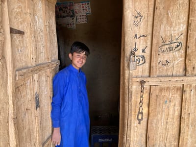 The image depicts a young man standing in the doorway of a building. He is wearing a blue long-sleeved shirt. The door is made of wood and is partially open. The door has a lock and a chain attached to it. There are inscriptions on the door in Arabic script. The scene appears to be in Saranan, Pakistan, based on the provided coordinates. The surrounding environment suggests a rural setting, with the building constructed of mud brick.  The young man is looking at the camera, while the interior of the building behind him is obscured from view.