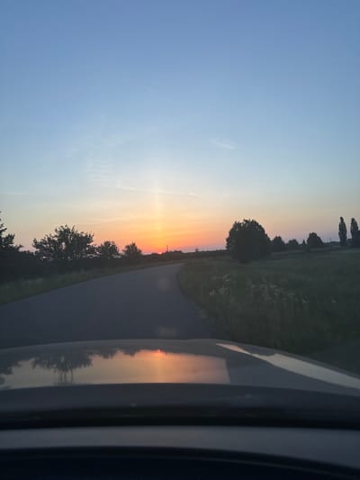 The image captures a rural scene at dusk, likely on a road in or near Dovhe, Ukraine. The sky is a gradient of pale blue at the top, transitioning to soft oranges and yellows near the horizon, indicating sunset. A distinct sun pillar effect is visible, a vertical shaft of light extending upwards from the setting sun. The sun itself is a small, bright orange disc partially obscured by a thin atmospheric layer.
A paved road curves gently through the landscape, leading the viewer's eye towards the sunset. To the left of the road, silhouetted trees and foliage form a dark band against the sky. To the right, a grassy field with some wildflowers stretches towards the horizon, dotted with more trees, including a few tall, slender poplars in the distance. The foreground shows the reflection of the sunset and surrounding trees on the hood of a vehicle, suggesting the photo was taken from inside a car. The overall impression is one of peaceful, natural beauty in a rural setting.