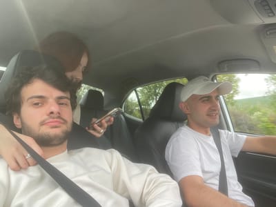 Two young men are inside a car. One on the left wears a white T-shirt and a white baseball cap. The other man on the right wears a white sweater and has a black seatbelt strap over his body. He is holding a mobile phone in his hand. The car appears to be in motion, as indicated by the blurred scenery visible through the window. The location cannot be determined based on the image.