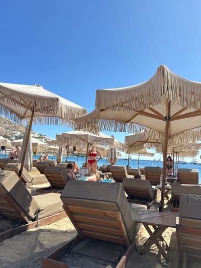The image depicts Ornos Beach in Ornos, Greece, on a clear, sunny day. The scene is dominated by rows of off-white, octagonal beach umbrellas featuring macrame-style fringed edges, supported by wooden poles, casting partial shadows on the light-colored sand. Beneath these umbrellas are numerous dark-cushioned wooden lounge chairs/sunbeds, some with adjustable backrests. Small, foldable wooden side tables are positioned between some sunbeds.
In the midground, several individuals are present. A person in a red two-piece swimsuit stands facing left near a sunbed. To their left, another person, possibly a child or young adult, is seated on a sunbed, facing right. Further right, closer to the water's edge, a person in a dark swimsuit stands. Additional individuals are visible in the background, seated on sunbeds or near the water.
The background features the calm, blue waters of the bay, dotted with numerous white boats and yachts anchored at varying distances. Beyond the bay, distant hills are visible on the left horizon, sparsely populated with white structures. The sky is clear and bright blue.