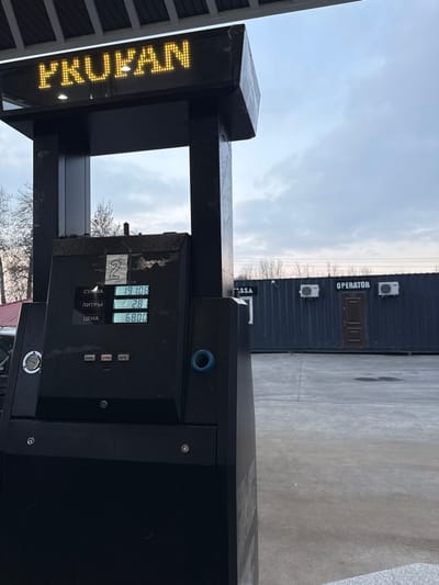 The image depicts a static scene at a gas station in Argin, Uzbekistan. In the foreground, a black fuel pump is prominently positioned. An LED sign at the top of the pump displays the Cyrillic word "ФРУВАН" (FRUVAN) in yellow letters. The pump's digital screen shows "СУММА" (SUM) as 19.10, "ЛИТРЫ" (LITERS) as 2.8, and "ЦЕНА" (PRICE) as 68.00, indicating a fuel transaction in progress or completed. A handwritten number "2" is taped onto the upper part of the display panel. A blue circular ring, likely a nozzle holster, is visible on the right side of the pump. The ground is paved with concrete or asphalt. In the midground, a dark grey or black corrugated metal container building serves as a station facility, featuring a brown door and two white exterior air conditioning units. White Cyrillic text on this building identifies sections as "KASSA" (CASHIER) and "OPERATOR." The background consists of bare trees beneath an overcast grey sky. No individuals, vehicles, or active fueling operations are visible.