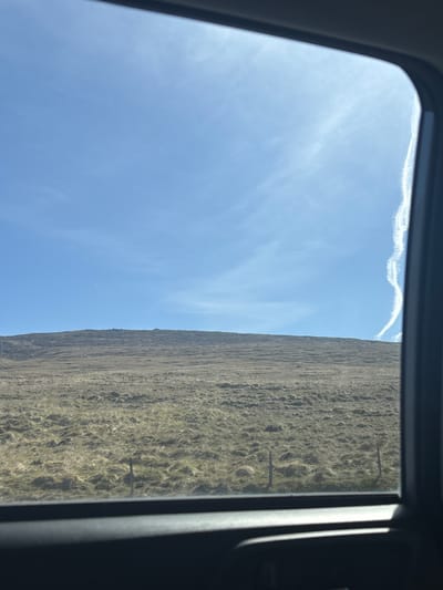 The image shows a view through a car window towards a vast, treeless, grassy hillside under a bright blue sky with a wispy contrail on the right side. The ground is covered in dry, golden-brown grass, with some patches of darker earth visible. A simple fence made of wooden posts and wire runs across the foreground of the hillside. The window frame itself is visible in the foreground, obscuring the lower part of the scene. The overall impression is of a remote, open landscape, characteristic of the rural areas around Borðeyri, Iceland. There are no people or animals visible in the frame. The time of day appears to be daytime, with strong sunlight casting shadows.