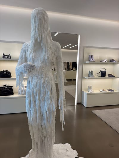 The image depicts a fashion boutique in Reggello, Italy. In the foreground, a life-sized mannequin is covered in a white, dripping material, resembling melted wax. The mannequin is positioned in front of shelves displaying handbags and shoes. The shelves are illuminated with strip lighting, and the overall aesthetic is minimalist and modern. In the background, a large mirror reflects more of the store's interior, including racks of clothing. A light-colored rug is visible on the dark brown floor.