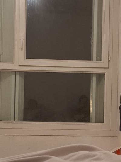 A close-up depicts a white window frame with two panes. The left pane is opaque, while the right pane reveals a blurred, ghostly figure peering in. The figure appears to have dark, round eyes and a nose. The window has a simple, modern design. A white shirt sleeve is partially visible on the right side of the frame. The wall surrounding the window is painted a neutral color.
