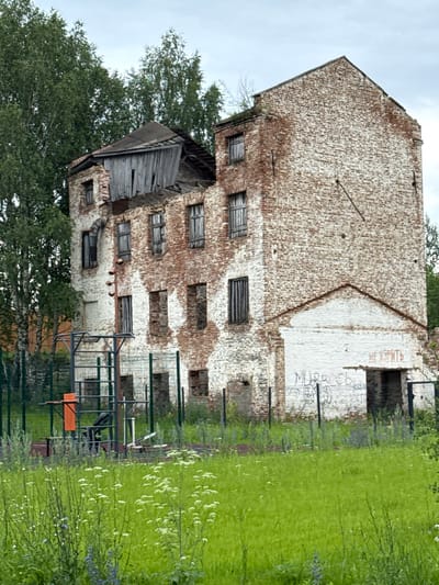 The image shows a dilapidated, multi-story brick building in Verkhotorie, Russia. The building is in a state of severe disrepair, with a partially collapsed roof and numerous broken windows. The brickwork is weathered and stained, with some areas whitewashed. Graffiti is visible on the lower exterior walls. In the foreground, there is a chain-link fence surrounding an outdoor exercise area with various pieces of gym equipment, including pull-up bars and a weight machine. The ground is covered in lush green grass and wildflowers. Tall trees are visible behind the building, and the sky is overcast.