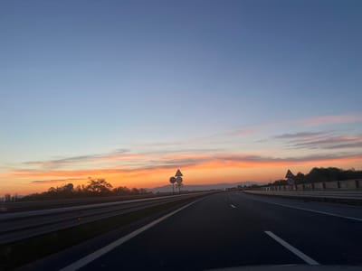 A highway runs through a rural landscape as the sun sets over Malo Buchino, Bulgaria. The sky is a gradient of blue, transitioning to orange and pink hues as the sun dips below the horizon. Wispy clouds are scattered across the sky, catching the last rays of sunlight. On the left side of the highway, a metal guardrail separates the road from a tree-lined embankment. Further in the distance, a large, dish-shaped structure is visible atop a hill, surrounded by trees. On the right side of the highway, another guardrail runs parallel to the road, with more trees and vegetation beyond. Several vehicles are visible on the highway in the distance. Traffic signs are posted along the side of the road, including one indicating a speed limit of 70. The overall scene depicts a peaceful evening drive through the Bulgarian countryside.