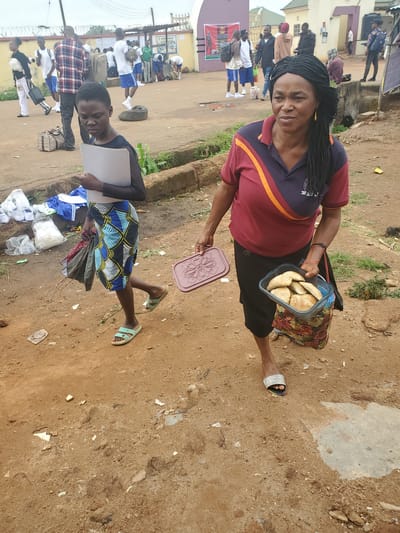 In a dusty, outdoor environment in Bahit, Nigeria, a young girl walks in the foreground, holding a grey folder and wearing a dark top with sheer sleeves and a colorful patterned skirt. She is followed closely by an adult woman carrying a brown plastic container filled with what appear to be pastries in one hand and a patterned bag in the other. The woman wears a maroon and navy polo shirt. In the background, other individuals are visible, some in athletic attire, suggesting a school or sporting event. Several bags and scattered white items, possibly clothing or supplies, are on the ground near the girl. The ground is a mix of dirt and gravel, with some concrete structures and vegetation visible. The overall scene depicts a casual, possibly transitional moment, with elements of daily life and community present.