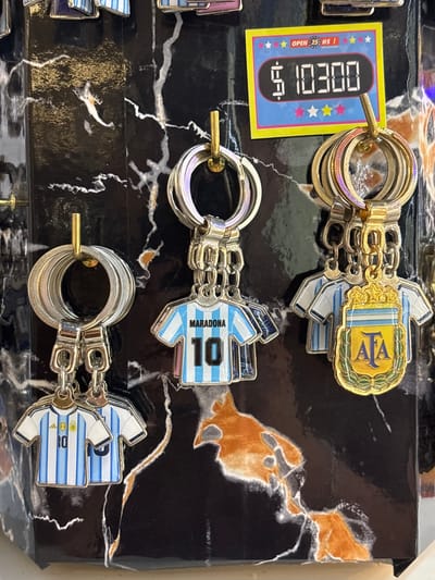 The image displays a collection of keychains with a football theme, specifically related to the Argentinian national team. The keychains are arranged in rows on a dark, glossy surface with a marbled pattern.

The top keychain features the AFA (Asociación del Fútbol Argentino) logo, a stylized shield with the letters "AFA" in gold on a blue and white background, bordered by a golden laurel wreath. Below it, several identical miniature football jerseys are attached. These jerseys are designed with the iconic light blue and white stripes of the Argentinian national team and have the number "10" on the back.

The second row of keychains also features miniature football jerseys, again in the Argentinian national team's colors with the number "10" and the name "MARADONA" visible on the back.

The bottom row shows another set of similar jerseys, also numbered "10" and representing the Argentinian national team.

To the left of the top keychain, a small yellow and blue price tag is visible. It has a digital display showing "00:29", suggesting a price in Argentinian Pesos or possibly a time-based sale. Text on the tag reads "OPEN 24 HS".

The overall scene appears to be a retail display, likely within a gift shop or souvenir store at Aeropuerto Internacional Ezeiza in Argentina, given the context provided. The lighting suggests an indoor environment, and there are no indications of weather or time of day beyond the "OPEN 24 HS" text. The objects are presented neatly, implying a commercial setting. There are no people visible in the image.