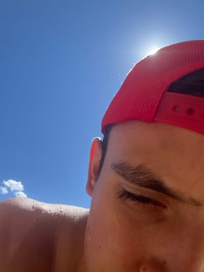 The image shows a close-up of a person wearing a red baseball cap, looking downwards against a bright blue sky with the sun shining. The person's face is partially visible, with an eye and eyebrow in focus. Their skin appears to be wet, with water droplets visible on their shoulder and upper arm. The background is dominated by the clear sky, with a small white cloud on the left side. The sun is positioned above and behind the cap, creating a bright glare and some lens flare. The image does not contain any information that allows for its location to be identified in Delchevo, North Macedonia.
