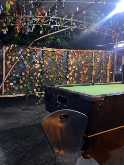 The image depicts an outdoor or semi-enclosed commercial space during nighttime, located in Igbessa, Nigeria. In the immediate foreground, a dark, possibly metallic, chair with a cutout back is partially visible on the right. A billiards table with a dark frame and a green felt playing surface occupies the mid-ground right. Behind the billiards table, a dark-colored wall or partition is visibly segmented by horizontal and vertical supports, covered extensively with artificial leaves in varied hues of green, orange, blue, and yellow. An overhead metal truss structure, similarly adorned with artificial foliage and strung with illuminated white fairy lights, spans the upper portion of the scene. The ground is paved with dark, patterned tiles. Beyond the decorated wall, natural dark green foliage is visible against the night sky. A faint, indistinct human silhouette is discernible in the far background to the right, adjacent to a partially visible sign containing the letters "GIFT." No active human interaction or specific actions are observed.