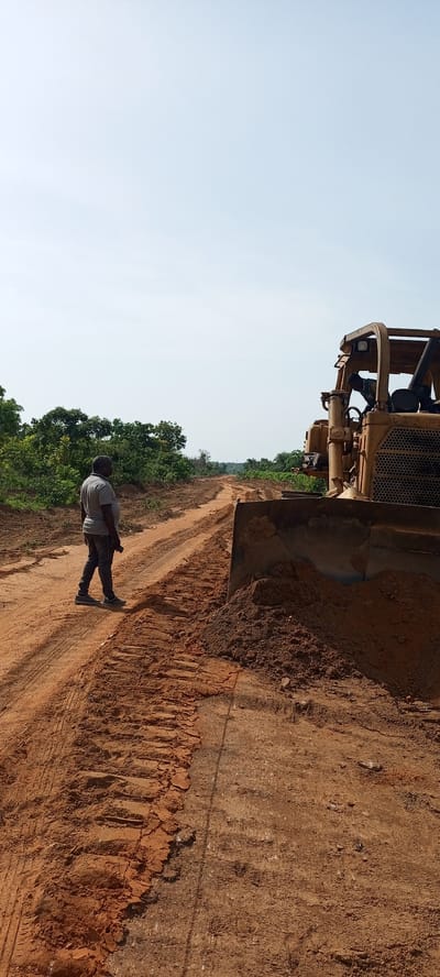 The image captures a scene of land clearing or road construction in a rural area of Otunade, Nigeria. A wide, unpaved road of reddish-brown earth, marked with tire and track impressions, extends from the foreground into the distance.

In the left mid-ground, a man stands on the dirt road, facing away from the viewer and looking towards the heavy machinery. He is wearing a grey short-sleeved shirt, dark pants, and sneakers, appearing to supervise or observe the work.

Dominating the right side of the frame is a large, yellow bulldozer, with its front blade lowered and pushing a substantial pile of freshly excavated soil. A person can be indistinctly seen inside the open cabin of the bulldozer, operating the machinery.

The background features abundant green foliage, consisting of trees and bushes, lining both sides of the developing road. The sky above is clear and light blue, indicating a bright, sunny day. The scene conveys an ongoing infrastructure project in a natural, undeveloped setting. No discernible text is visible in the image.