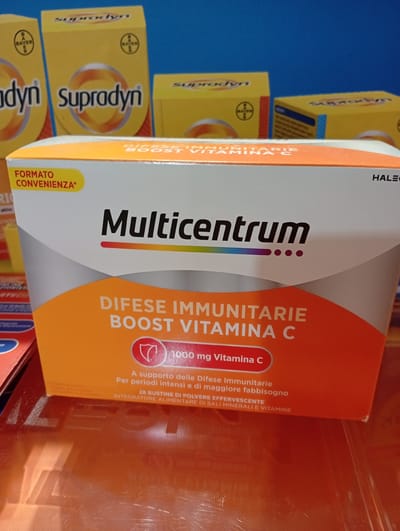 The image shows a close-up of a box of Multicentrum vitamins. The box is primarily white and orange, with the brand name "Multicentrum" prominently displayed in black text. Below the brand name, in smaller text, it says "DIFESE IMMUNITARIE BOOST VITAMINA C," indicating the product is for immune defense and contains Vitamin C. There is also a red circular emblem with a shield icon and the text "1000 mg Vitamina C" within it. The box is angled slightly, revealing a portion of the side with "FORMATO CONVENIENZA*" in yellow text. In the background, out of focus, are several yellow boxes of "Supradyn" vitamins, also with the Bayer logo. The scene appears to be an indoor display, possibly in a pharmacy or retail store. There is no information available in the image that would place this scene in Civitanova Marche, Italy.