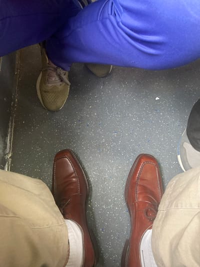 The image displays a top-down view of the lower extremities of three individuals within an interior space, reportedly located in Partido de Lanús, Argentina. The floor is a dark grey, speckled surface with scattered lighter flecks. In the foreground, a person is seen wearing light brown khaki trousers, white socks, and a pair of reddish-brown leather square-toed dress shoes. The right shoe features a subtle circular emblem on its upper. Towards the upper left, another individual's legs are crossed, with royal blue trousers and one dark olive green athletic shoe with a white sole visible. A third person's grey and black athletic shoe is partially visible on the far right of the frame, adjacent to the right foot of the person in khaki trousers. A dark vertical line marks the left boundary of the visible floor area. No specific actions or interactions between the individuals are discernible beyond their proximity.