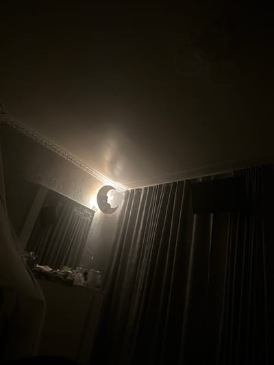 The image depicts an indoor scene with dim lighting, possibly a bedroom or nursery. A crescent moon-shaped night light, featuring a silhouette of a bear and a star, is illuminated on the wall, casting a soft glow. The wall has a textured finish and is adorned with decorative molding along the ceiling. To the left, a surface is cluttered with various items, including bottles and other unclear objects. Heavy, dark curtains hang on the right side of the frame, obscuring the view of the rest of the room. A ceiling fan is faintly visible at the top of the image. The overall impression is one of a dimly lit, perhaps child-friendly, environment. There are no visible people or animals in the image. The location in Prokhladny, Russia is not discernible from the image content.