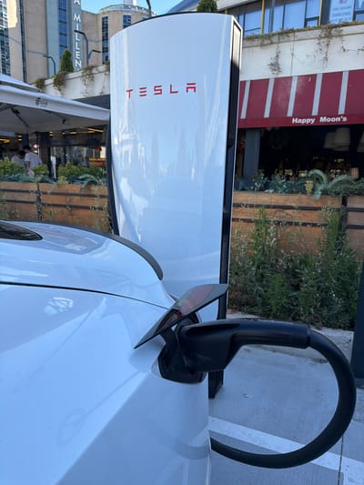 In this photograph, a white Tesla charging station stands prominently, its sleek design contrasting with the surrounding environment. A black charging cable is plugged into a white Tesla electric vehicle, indicating that the car is actively charging. The background reveals a street scene with a building displaying a sign that reads "Happy Moon's." Adjacent to it, another building features a sign with "MILLEN" partially visible. The scene appears to be set in an urban area, possibly a commercial district, given the presence of businesses and charging infrastructure. The lighting suggests it is daytime, and the weather seems clear. The composition focuses on the interaction between the electric vehicle and the charging station, hinting at the growing adoption of electric mobility. The overall impression is one of modern convenience and technological advancement.