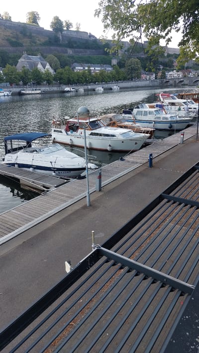The image shows a waterfront scene in Namur, Belgium, with several boats docked along a wooden pier. The boats appear to be recreational vessels, with varying sizes and styles. Some of the boats have cabins and windows, while others are more open. The pier is made of wood planks and has metal railings and bollards for securing the boats.

In the background, on the left side of the image, a steep, tree-covered hill rises. On top of the hill, there are structures that appear to be part of a historic fortification or castle. Further down the hill and along the waterfront, there are buildings that look like houses or commercial establishments, interspersed with trees. The water is calm and reflects the sunlight, indicating it's likely daytime.

The overall impression is a peaceful, scenic location, possibly a harbor or marina. There are no people clearly visible in the image, suggesting the activity is centered around the docked boats. The lighting suggests it could be late morning or early afternoon, with bright sunlight casting shadows. The presence of the fortifications in the background provides a strong geographical clue to the location being Namur, Belgium, known for its citadel overlooking the confluence of the Sambre and Meuse rivers. Some of the boats have visible text, such as "Dora" and "B43247" on one of the vessels. The scene is serene, with the water, boats, and historical architecture creating a picturesque setting.