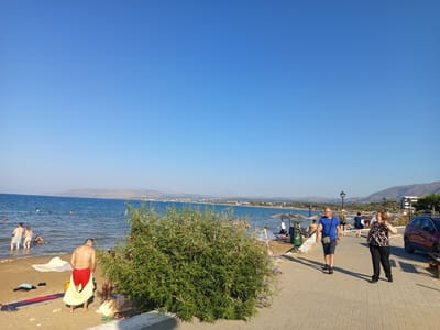This image captures a sunny day at the beach in Georgioupoli, Greece. The scene depicts a section of the coastline with the blue sea meeting a sandy beach. On the beach, several people are visible, some wading in the water, while others are on the sand, with towels and beach accessories spread out. A man in red shorts is seen adjusting his attire, and a couple stands in the shallow water. To the right, a paved walkway runs alongside the beach, with several people strolling along it. Two individuals, a man in a blue t-shirt and a woman in a patterned top, are prominently featured walking on the path. Beach umbrellas and lounge chairs are set up further down the beach, indicating a popular spot for relaxation. In the background, lush green foliage is prominent, obscuring some of the view. The overall environment is bright and clear, with a panoramic view of the sea and distant hills. A red car is parked on the right side of the walkway.