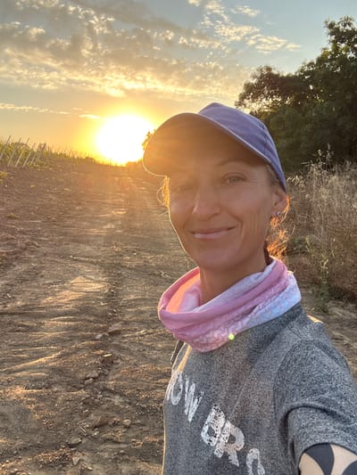 A woman stands in a selfie, looking at the camera with a smile. She wears a blue baseball cap, a pink and white patterned neck gaiter, and a grey sweatshirt with white lettering. The background is a dirt path leading uphill, with vineyards on the left and dry brush and trees on the right. The sun is setting in the distance, casting a warm glow over the scene. The location is stated to be in the Gurzufsky settlement council, Ukraine.