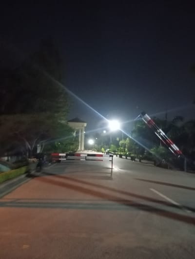 The image captures a nighttime scene at a guarded entrance in Rawalpindi, Pakistan. A paved road stretches into the distance, illuminated by streetlights. A red and white striped barrier arm blocks the road, with a matching barrier on the left side of the frame in a closed position. A guard is visible on the left, near the barrier. In the distance, a building with white columns is visible. Vegetation lines the sides of the road, and the sky is dark.