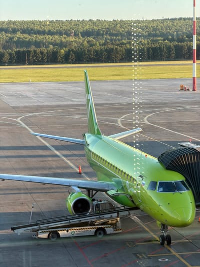 The image shows a green S7 Airlines Airbus A320neo aircraft parked at a gate at the Емельяново Airport (KJA) in Krasnoyarsk, Russia. The aircraft is connected to a jet bridge. A ground support vehicle is parked alongside the aircraft. A runway with lights and a grassy area bordered by trees are visible in the background.