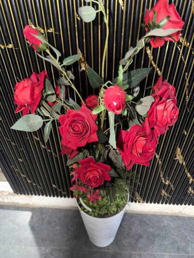 This media file features a close-up, overhead view of a bouquet of artificial red roses arranged in a white cylindrical vase. The vase is placed on a surface covered with black horizontal blinds that have thin gold lines running through them. The background transitions from the blinds to a mottled grey surface, suggesting an indoor setting possibly near a wall. The lighting is natural and appears to be from above, casting soft shadows. The roses are in various stages of bloom, with some fully open and others still budding. The leaves are a muted green, and the stems are visible, wrapped in green material. The overall impression is one of artificial elegance, with the bright red of the roses contrasting with the dark blinds and white vase. There is no visible text in the media file. The setting does not contain any identifiable landmarks or cultural elements specific to Khumsan, Uzbekistan, other than the provided context.