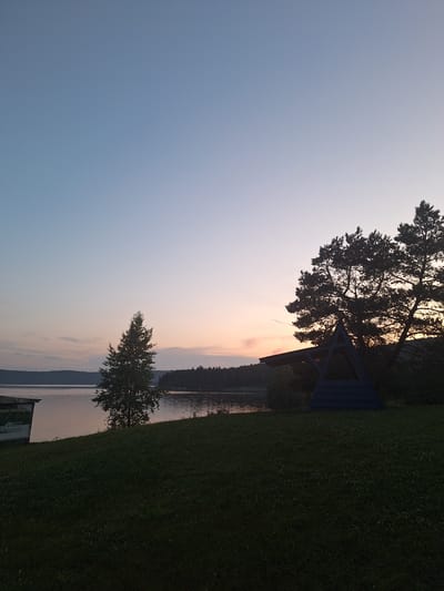 The image depicts a serene evening landscape with a clear sky transitioning from light blue to soft orange hues near the horizon. A calm body of water, likely a lake or river, stretches across the mid-ground, bordered by a tree-lined shore in the distance. In the foreground, a neatly mowed grassy hill slopes downwards towards the water. A single, slender tree stands prominently on the left side of the hill, while on the right, a dark silhouette of pine trees frames a triangular wooden structure, possibly a gazebo or shelter, illuminated by dim light. A small, weathered building with visible signage is partially visible on the far left, near the water's edge. The overall scene suggests a tranquil, natural setting at dusk, potentially in the vicinity of Атамановка, Russia, given the context provided.