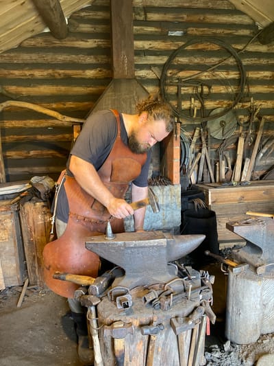 In Patanevshchina, Russia, a blacksmith with a beard and curly hair is working in his shop. He is wearing a leather apron over a dark t-shirt and is focused on his work, holding a hammer over an anvil. The shop is rustic, with log walls and a wooden ceiling. Various blacksmithing tools, including hammers, tongs, and punches, are scattered around the anvil and on wooden blocks. In the background, coils of wire, more tools, and a charcoal bin are visible. The overall impression is one of a traditional, working blacksmith's forge.