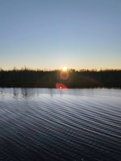 The image depicts a body of water with gentle, linear ripples reflecting the sky and a dark treeline silhouetted against the setting sun. The sun is low on the horizon, creating a bright glare and lens flare. The sky is a gradient of light blue to pale orange. The location is likely near Kurdiug, Russia, based on the potential geographical features and landscape. No people or specific objects besides the water, trees, and sun are visible.
