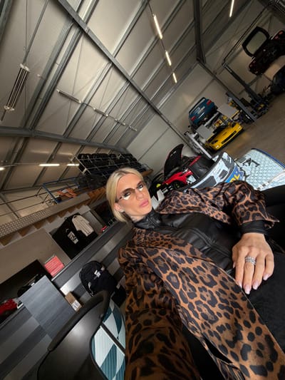The image depicts a female subject seated within an indoor automotive workshop located in Meuspath, Germany. The subject, a woman with blonde hair, is wearing small, rimless sunglasses, a black top, and a prominent brown leopard-print jacket or scarf. She is positioned towards the left of the frame, looking slightly upward and to her left with a subtle facial expression. Visible tattoos are on her neck/chest area, and she has multiple rings on her left hand.
The environment is a large industrial space with a high, white-paneled ceiling supported by metal beams, featuring multiple linear fluorescent light fixtures and infrared heaters. The floor appears to be concrete or epoxy. In the background, numerous vehicles are present. Directly behind the subject to her right, a red sports car with an open hood is visible, next to a white/silver race-prepared vehicle. Further into the background, a blue car is elevated on a hydraulic lift, with a yellow sports car positioned beneath it on another lift. A dark-colored vehicle is on an even higher lift in the far background. Along the left side of the background, racks of multiple tires are stacked. A desk or counter area with a red binder and folded white shirts is visible to the left of the seated subject. A black backpack rests near the desk. The subject is seated in a black chair, part of which is visible in the lower left corner of the frame, with a patterned surface (teal and white checkered) also partially visible.