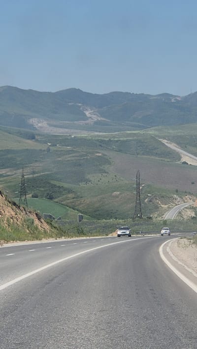 A wide, asphalt road curves through a hilly, green landscape under a clear blue sky. Two cars are traveling on the road, one silver sedan in the left lane and a white SUV in the right lane. The landscape is characterized by rolling green hills, some of which appear to be cultivated with varying shades of green. In the distance, more hills are visible, with a dusty, lighter brown area on one of the slopes suggesting some sort of excavation or construction. Two tall, lattice-style power line towers are present in the midground, one to the left of the road and another further back to the right. The road is marked with white lines, and the shoulders on either side are composed of gravel and sparse vegetation. The scene is natural and appears to be taken from a moving vehicle on the road.