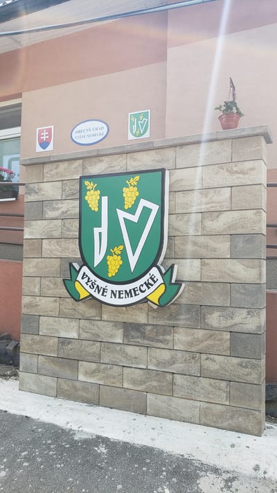 This media file depicts a street-level view of a building facade adorned with several signs and a decorative element. The main focus is a large, green, shield-shaped emblem with yellow grapes and a stylized "14" within it. The emblem is outlined in black and has a yellow and green wavy border. Text on a white banner accompanying the emblem reads "VYŠNÉ NEMECKÉ".
To the left of the emblem, on a beige and light brown striped wall, are three smaller signs. The topmost is a small green and white square with the same "14" symbol. Below that is a blue oval with white text that reads "OBECNÝ ÚRAD VYŠNÉ NEMECKÉ". The bottom sign is a square with the Slovak flag. A small potted plant with green leaves is visible on a ledge above these signs.
The wall is constructed with large, rectangular light grey and beige stone blocks. To the right of the wall, a textured grey concrete surface slopes down and meets dark asphalt. The lighting suggests it is daytime, possibly sunny, with a strong glare visible on the left side of the building wall. The overall impression is one of signage and architectural detail for a municipal building or landmark in Vyšné Nemecké, Slovakia. No people are visible in the frame.